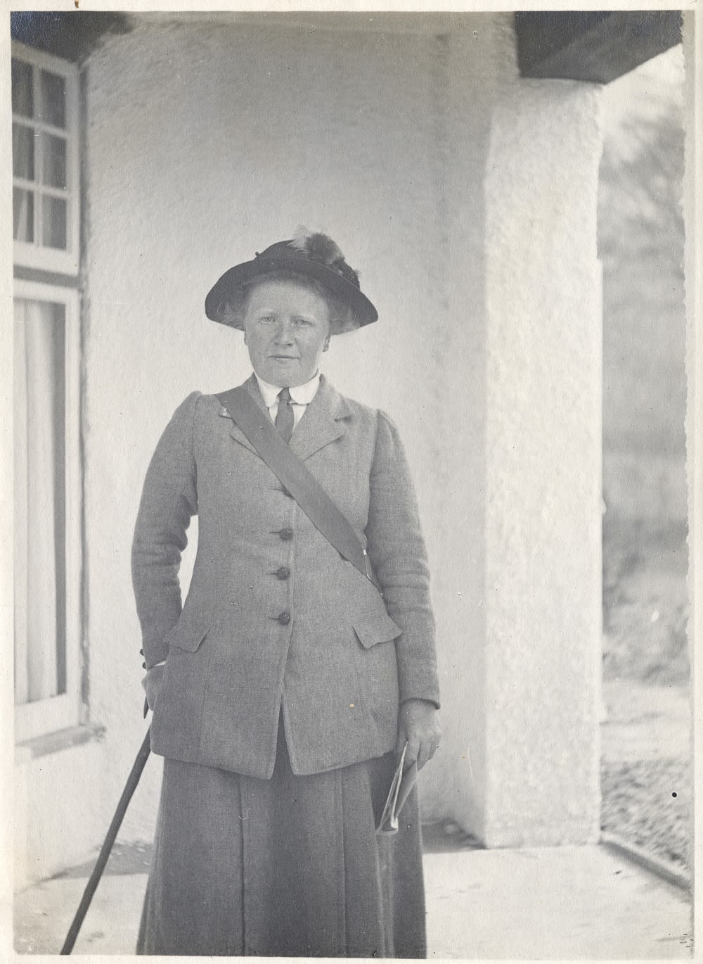 Photograph of a women in tweed clothing, wearing a hat and holding a cane and notebook. She has a satchel across her shoulder.