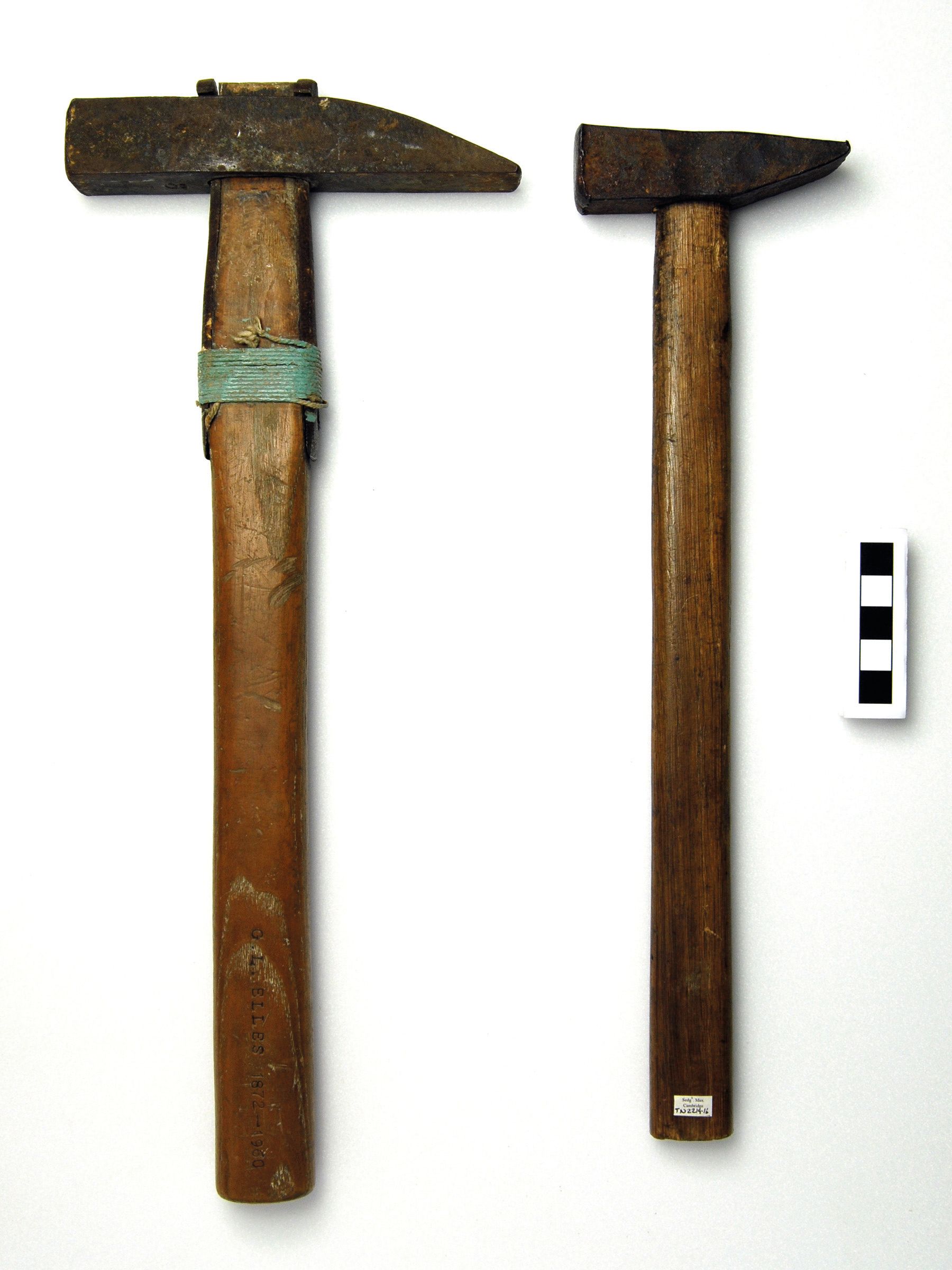 Photograph of 2 geological hammers with scale. One has a larger head than the other. The smaller one has a label stuck on the bottom. The larger one is enscribed with the name of the owner.