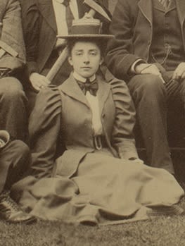 Formal photograph of a woman seated outside. She is wearing a hat and bow tie. A lady behind her is holding a large geological hammer.