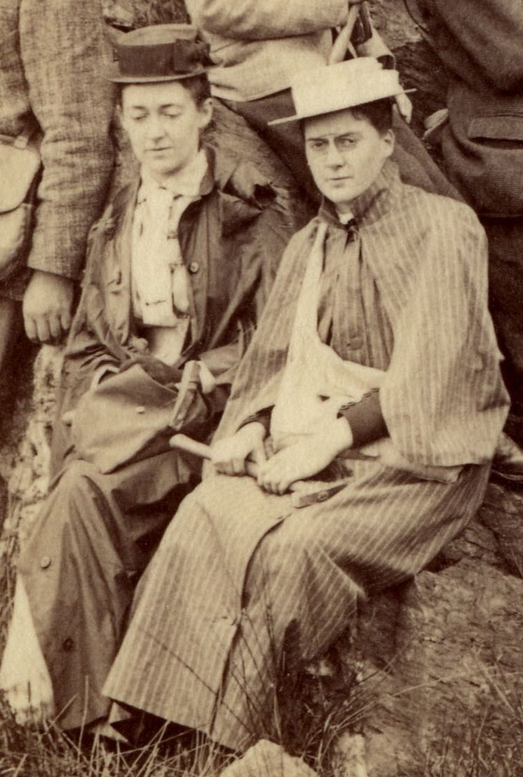 Photograph of two women sitting on rocks. They are both wearing hats and have bags around their shoulders. One is holding a geological hammer.
