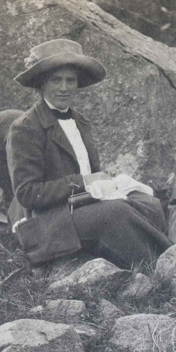 Photograph of woman outside eating lunch. She is wearing a large hat. She is surrounded by rocks.