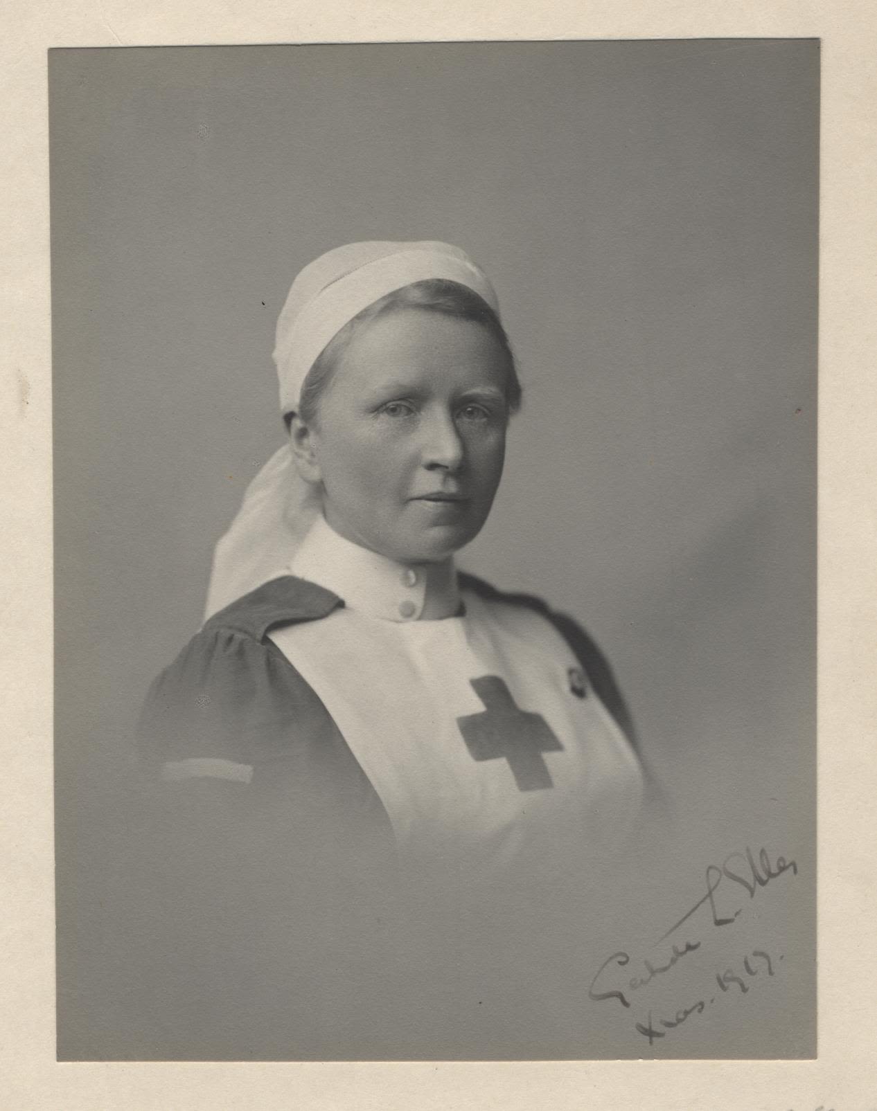 Photograph of woman in Red Cross nurses uniform. The front of the pinifore has a red cross on it. The photograph is signed by the sitter.