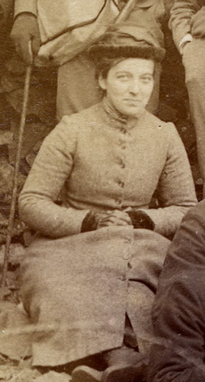 Photograph of women seated outside. She is wearing a long coat and a hat, with her hands clasped.