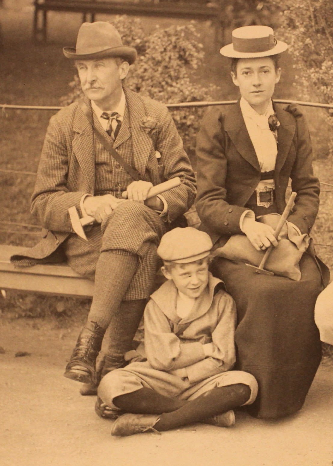 2 adults are seating on a bench wearing hats and holding hammers. A child is seated in front of them, with his arms crossed and wearing a hat.